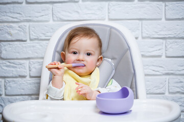 A small child sits on a highchair and eats food from a plate with a spoon. Baby silicone utensils for feeding babies