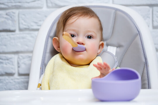 A small child sits on a highchair and eats food from a plate with a spoon. Baby silicone utensils for feeding babies
