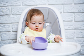 A small child sits on a highchair and eats food from a plate with a spoon. Baby silicone utensils for feeding babies