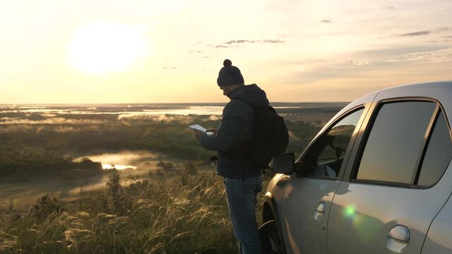 Driver-blogger tourist with tablet online on vacation. A tourist with a smartphone stopped at a campsite by car, looking at the sunset from mountain. A man traveling by car checks route in navigator