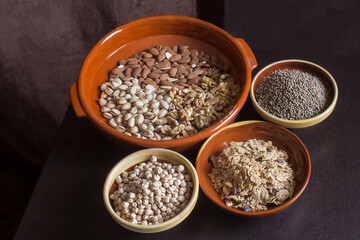 A large artisan clay pot with almonds, pistachios, and walnuts along with three bowls filled with lentils, chickpeas, and toasted flakes of rice, whole wheat, and chocolate on a wooden table.