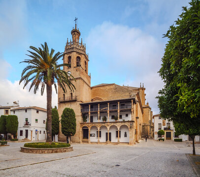 The historic Iglesia de Santa Maria la Mayor in Ronda, Spain, towering over Plaza de la Duquesa de Parcent with a large palm tree, traditional architecture, and a blue sky.