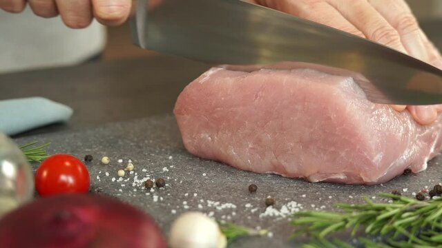 Chefs male hands cut fresh pork with large knife on background of onion, garlic, greens and spices. Food concept, cooking meat, loin, carbonate.