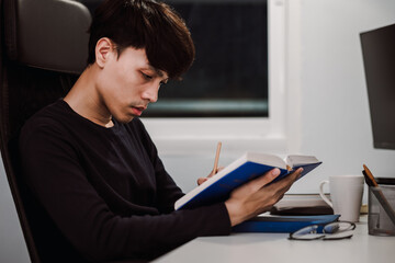 Young handsome asian man reading book and writing note at work desk late at night, Knowledge and learning concept.