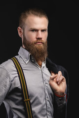 Portrait of a brutal attractive bearded Caucasian man in a shirt with suspenders and a jacket on his shoulder. Studio photography