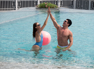 Beautiful happy couple in sunglasses with a beach ball enjoying their time together in the pool. Couple with sunglasses giving high five to each other in the pool..