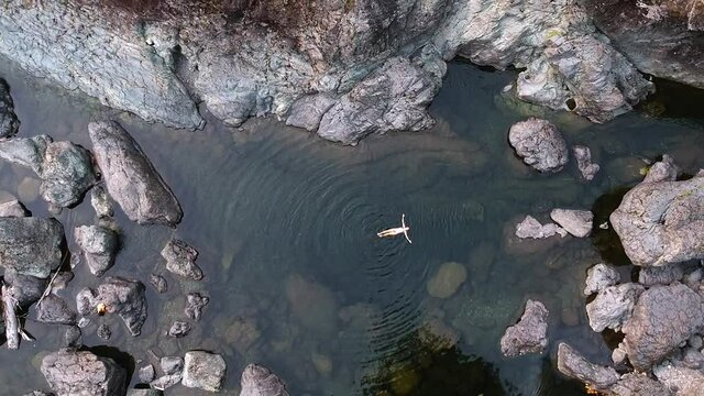 Top down bird's eye view drone shot of a girl in a bikini floating in crystal clear water in a rock pool located in the country side of British Columbia, Canada.