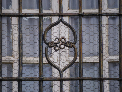 Close-up shot of an old window protected by a weathered wrought-iron security grille featuring a central decorative scroll emblem, layered over fine hexagonal wire mesh in front of a white multi-pane 