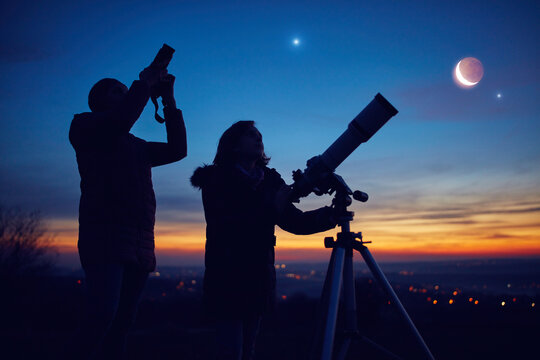Mother and daughter observing stars, planets, Moon and night sky with astronomical telescope.