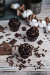 Several muffins or cupcakes with chocolate shaped cream at white table. Festive candle burns on a chocolate cake. There are chocolate chips scattered nearby