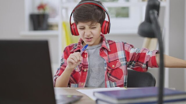 Portrait of excited joyful Middle Eastern boy in headphones singing and gesturing in slow motion sitting at table indoors. Careless schoolboy doing homework and enjoying music in earphones.