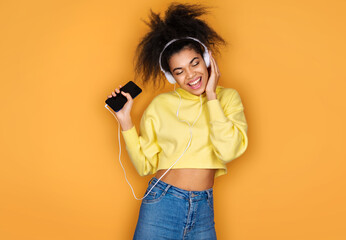 Young girl listens music to chill, holds smartphone and wears headphones on yellow background