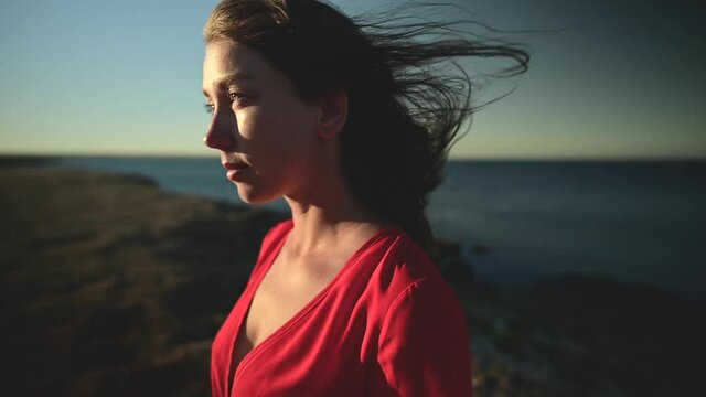 Attractive Caucasian young woman in a red dress by the sea looks towards the sun. Hair fluttering in the wind Close-up shallow depth of field slow motion.