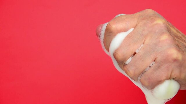 Old hands gripping a foamy sponge on a red background. Sponge in old hands. Soapy cleaning sponge in old hands. Disinfection and cleaning.Foam on a sponge in old hands. Cleaning and sanitation concept
