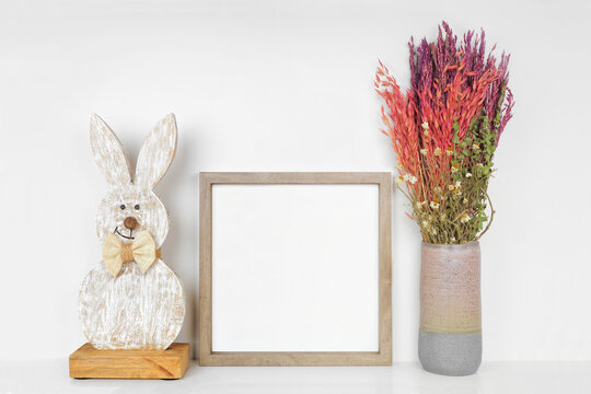 Mock up wood frame with Easter decor on a white shelf. Shabby chic wood bunny and vase of spring naturals. Square frame against a white wall. Copy space.