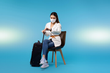 Pretty lady in a protective mask on a blue background, who sits on a chair with ready luggage and...