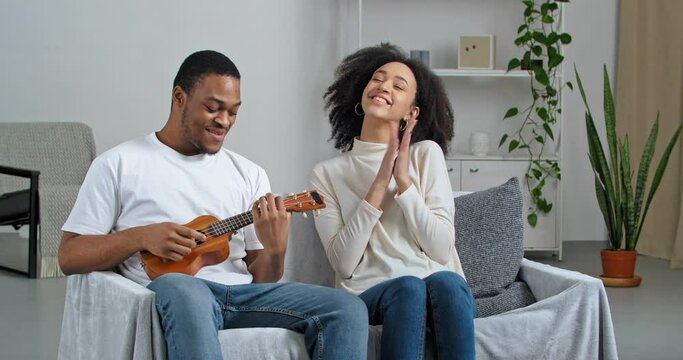 Young loving afro american guy husband boyfriend singing song playing small ukulele musical instrument for beloved beautiful woman curly girl wife girlfriend makes creative gift sitting at home couch