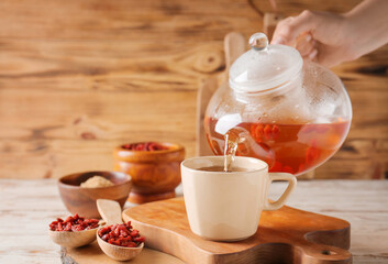 Woman pouring goji tea from teapot into cup on wooden background