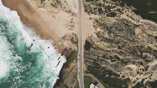 Aerial view of a desertic road driving next to a view of rough sea with crispy waves. Place situated in the south Portuguese wild coastline between the Atlantic ocean and the forest. Cascais, Portugal