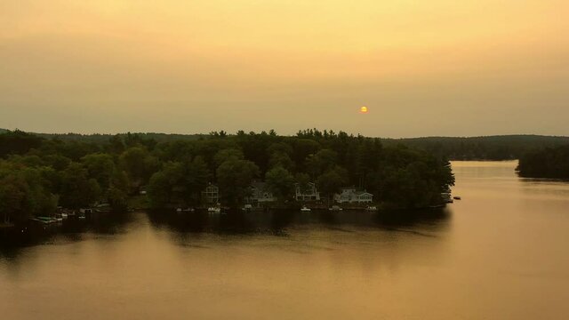 Aerial shot of houses amidst trees at lakeshore, drone ascending forward over lake against sky at sunset - Berkshire County, Massachusetts