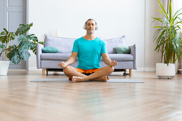 Photo of a middle aged man practicing yoga at home. He sits in the lotus position with headphones and listens to meditation music.