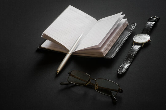 Glasses, a ballpoint pen, a leather-covered diary, and a wristwatch lie on a black table. The concept of planning a daily routine or filling out a personal diary.