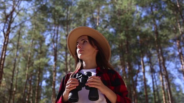 Woman in a hat holds binoculars while hiking in forest. Concept hike, tourism.