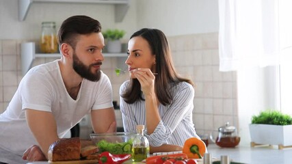 Happy young couple in good mood are preparing a fresh healthy vegan salad with many vegetables in the kitchen and man testing it from a girl's hands