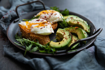 Delicious and delicious breakfast with poached egg on a slice of fried bread, avocado, arugula, cherry tomatoes and spices. Served in a cast-iron skillet, served with textiles on a black background . 