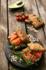 A delicious snack for lunch. Delicious sandwich with lightly salted salmon. With a fluffy bun, with arugula greens and sunflower seeds, with cherry tomatoes and avocado on the background. 