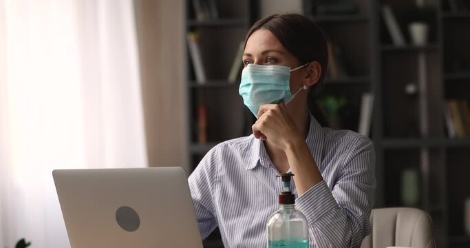 Focused busy young businesswoman in protective medical face mask working on computer in modern home office, has dispenser bottle with antibacterial gel on table for hands disinfection, hygiene concept