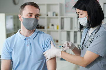 Female clinician or nurse in uniform and protective mask vaccinating young man