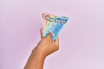 Hispanic hand holding canadian dollars banknotes over isolated pink background.
