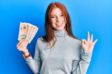 Young red head girl holding 10 united kingdom pounds banknotes doing ok sign with fingers, smiling friendly gesturing excellent symbol