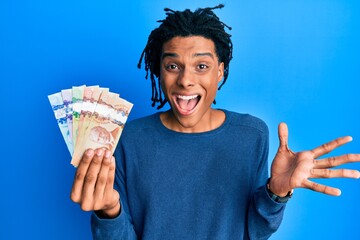 Young african american man holding canadian dollars celebrating achievement with happy smile and...