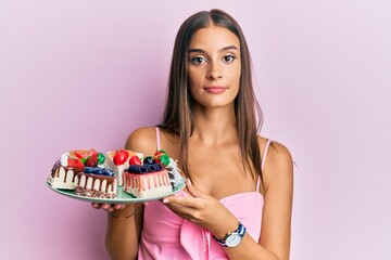 Young hispanic woman holding plate with cheesecake thinking attitude and sober expression looking self confident