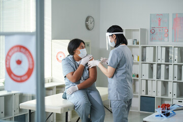 Young nurse in uniform and protective workwear making injection to her colleague