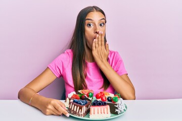 Young hispanic woman eating cheesecake sitting on the table covering mouth with hand, shocked and...