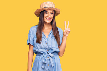 Young hispanic woman wearing summer hat smiling with happy face winking at the camera doing victory sign. number two.