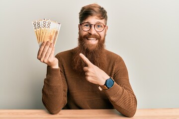 Young irish redhead man holding 500 norwegian krone banknotes sitting on the table smiling happy...