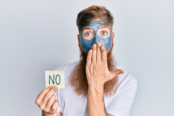 Young irish redhead man wearing facial mask holding no reminder covering mouth with hand, shocked...