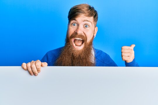 Young irish redhead man holding blank empty banner pointing thumb up to the side smiling happy with open mouth