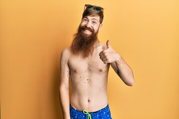Fototapeta premium Redhead man with long beard wearing swimsuit and sunglasses smiling happy and positive, thumb up doing excellent and approval sign