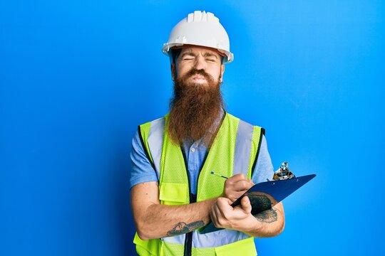 Redhead man with long beard wearing safety helmet holding clipboard puffing cheeks with funny face. mouth inflated with air, catching air.