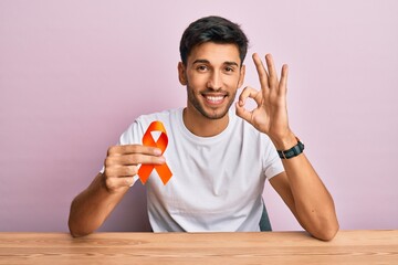 Young handsome man holding awareness orange ribbon doing ok sign with fingers, smiling friendly...