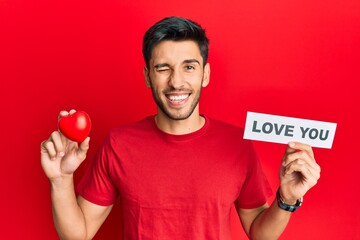 Young handsome man holding red heart and love you message winking looking at the camera with sexy...
