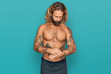 Handsome man with beard and long hair standing shirtless showing tattoos checking the time on wrist watch, relaxed and confident
