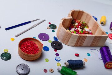 wooden heart shaped box of multicolored beads and buttons, awl, scissors, crochets and spool of threads for sewing and embroidery. Set of materials for handcraft, making of bijouterie and accessories.