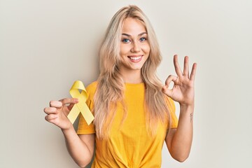 Beautiful caucasian blonde girl holding suicide prevention yellow ribbon doing ok sign with fingers, smiling friendly gesturing excellent symbol
