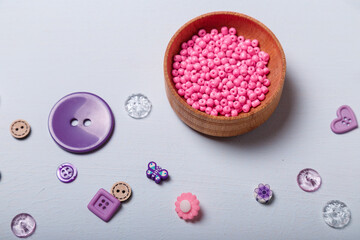 Wooden bowl of pink beads and purple multishaped buttons for sewing and embroidery on white background. Set of materials for handcraft, making of bijouterie and accessories. Copy space.